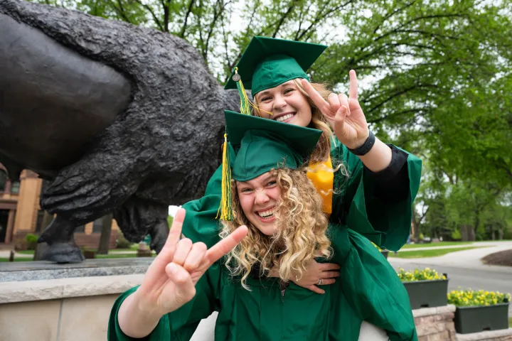 Two friends in commencement attire smiling in front of bison statue, holding up bison horns hand gesture