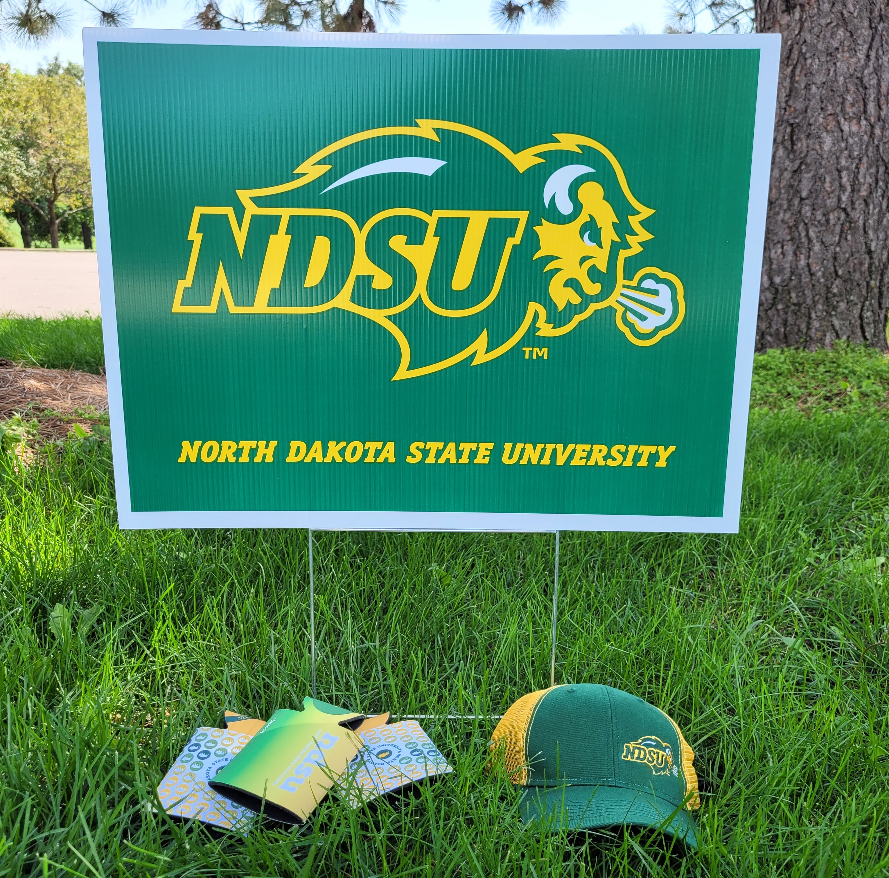 A Bison Athletics yard sign is posted in grass, with a Bison cap and can coozies on the ground in front of the sign.