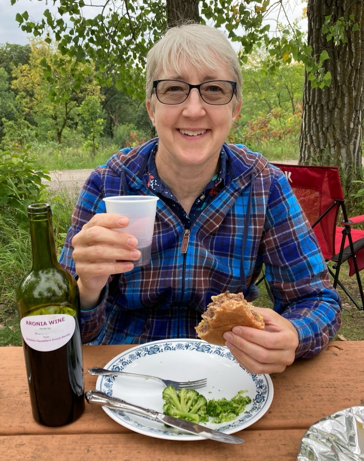 Kathy is sitting at a picnic table with food and some Aronia berry wine.