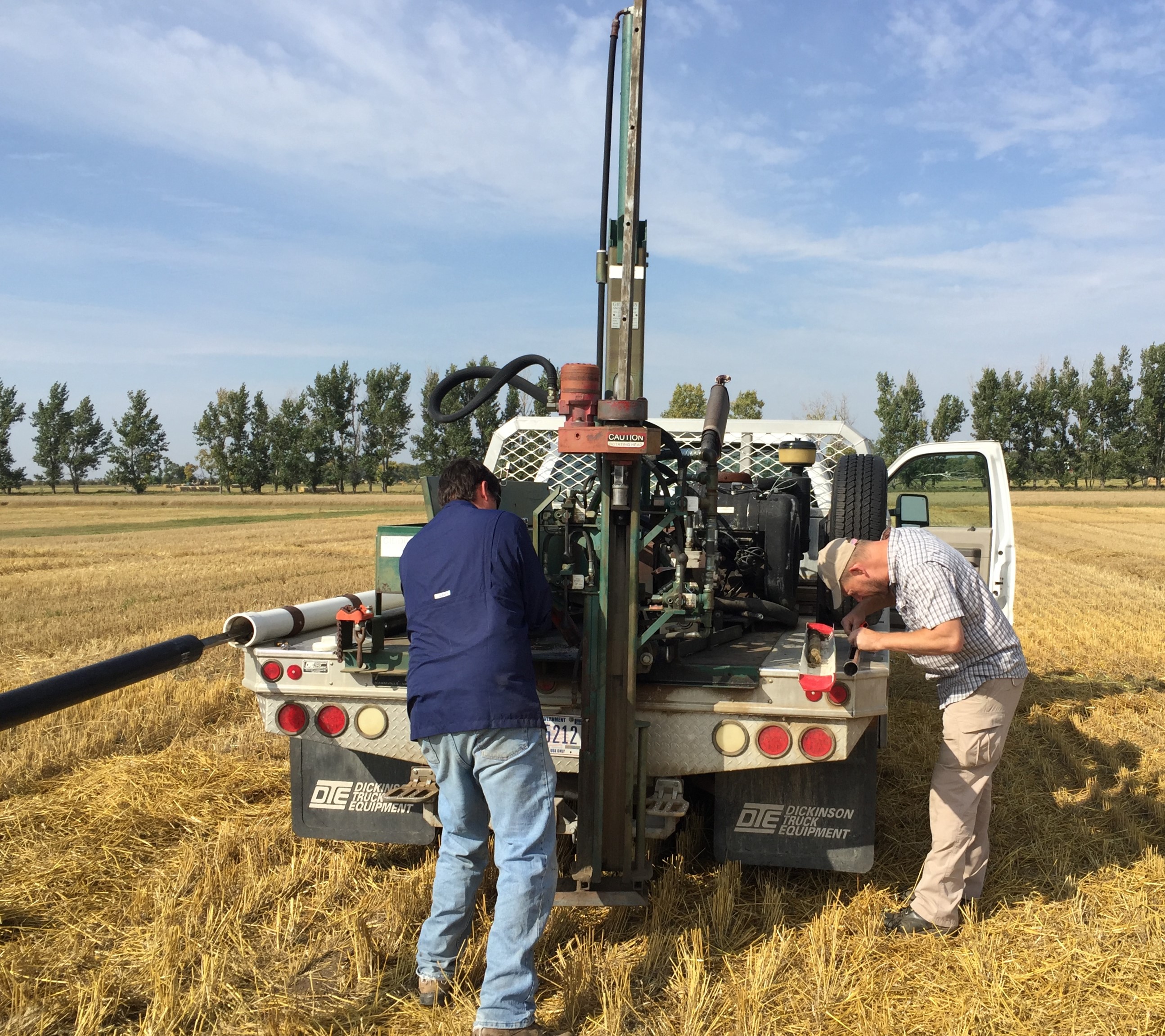 A hydralic soil probe is mounted on the back deck of a work pickup. Two men are taking soil samples from the stubble field.