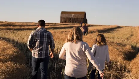 A family walks away from the camera through tall yellow grass toward an unpainted barn. Two kids are near the barn; parents and another child are closer.