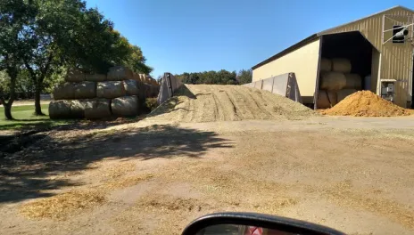 A large corn silage pile sits beside a yellow metal building, with a stack of hay bales and trees nearby.