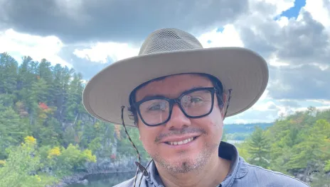 A man in a blue shirt with a tan hat stands in front of a river and trees, with a partly cloudy sky overhead.