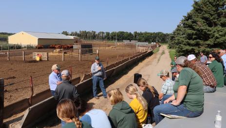 Two speakers lean on a feed bunk while talking to a group of people sitting on a flatbed trailer. A dirt feedlot with red cattle and a yellow polebarn are in the background.