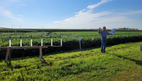 A spray boom display is parked in front of soybean plots. Michael Wunsch stands beside the display holding a sample handout.