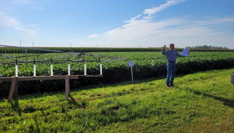 A spray boom display is parked in front of soybean plots. Michael Wunsch stands beside the display holding a sample handout.
