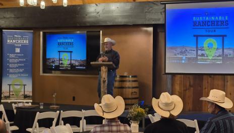 A lone man stands at a podium in front of an audience. Monitors behind him read "Sustanable Ranchers Tour."