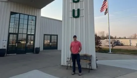 Juan Villeda at the North Central Research Extension Center. A young man is standing outside a large white building that is labeled, “North Central Research Extension Center” and “NDSU.” An American flag and a North Dakota state flag are flying from a pole in the background.