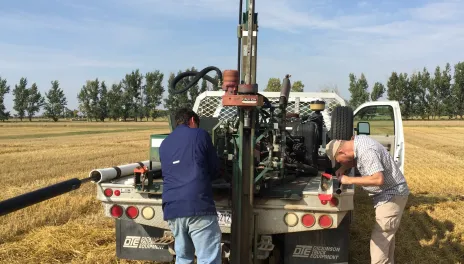 A hydralic soil probe is mounted on the back deck of a work pickup. Two men are taking soil samples from the stubble field.