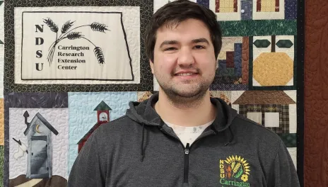 Agustin stands in front of a quilt with several blocks depicting agricultural themes.