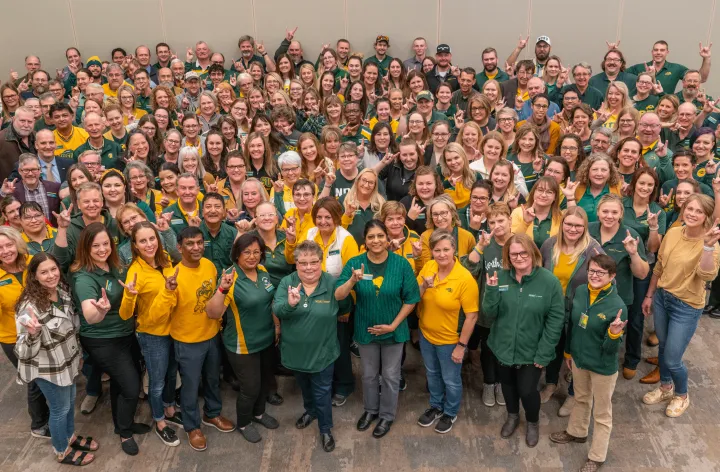 A large group of NDSU Extension personnel in green and gold attire pose making the "Bison Sign" with forefinger and pinkie raised.