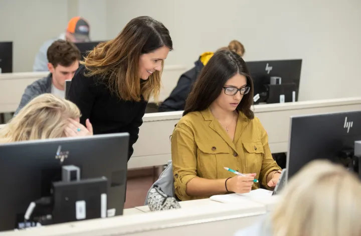 A student sits in front of a computer monitor as a teacher looks over their shoulder.