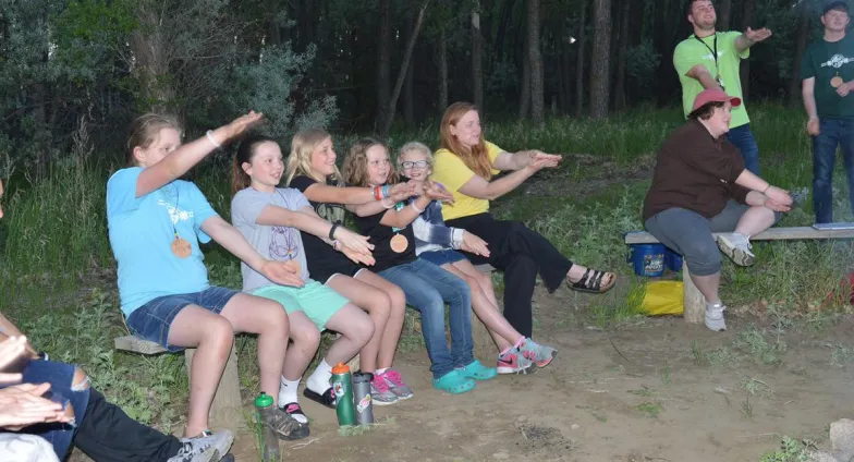 Youth and counselors in shorts and t-shirts gathered around a campfire at dusk.