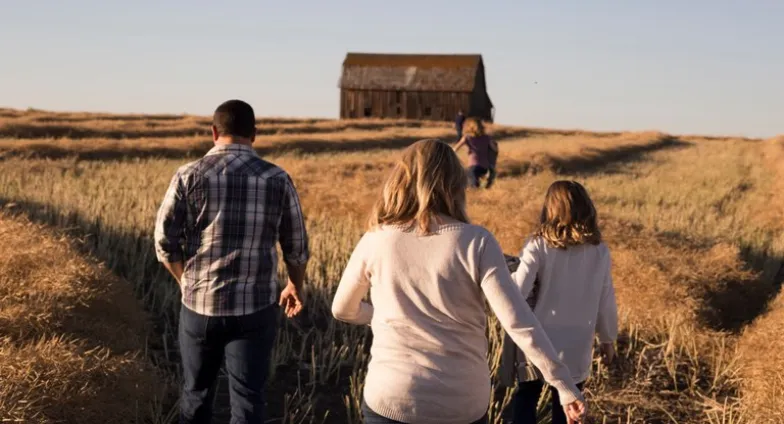 A family walks away from the camera through tall yellow grass toward an unpainted barn. Two kids are near the barn; parents and another child are closer.