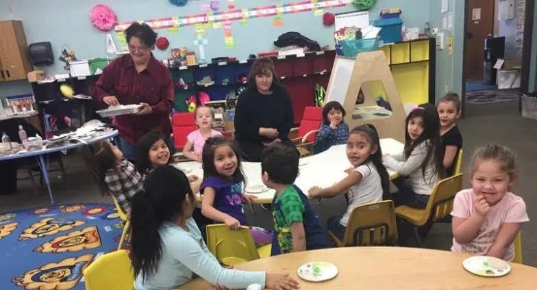 Twelve young kids sit around two round tables in a colorful classroom. One adult is with them while another passes out snacks.