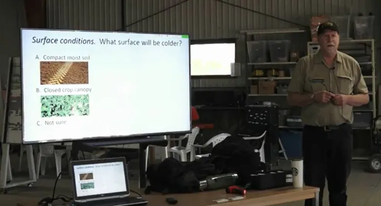 NDSU Pesticide Program specialist Andrew Thostenson, in a black cap and tan shirt, stands by a screen displaying, "Surface conditions: What will be colder?"