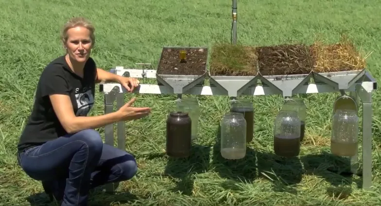 Soil health specialist Abbey Wick leans next to a rainfall simulator with four soil containers on top and clear bottles below.