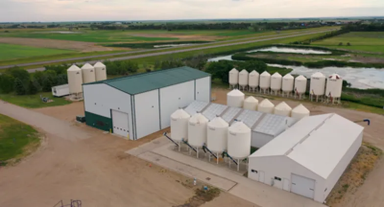 An aerial view shows two large metal buildings—one all white, the other white with a green roof—surrounded by several white silos.