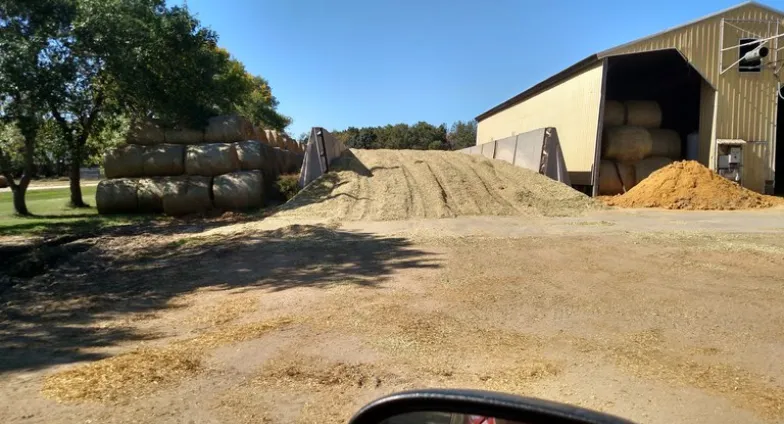 A large corn silage pile sits beside a yellow metal building, with a stack of hay bales and trees nearby.