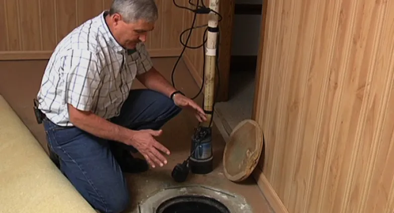 A man kneels beside a sump pump hole in the floor of a basement.