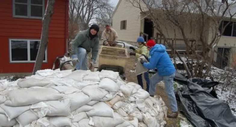 Four people in winter coats and hats fill white bags with sand and stack them into a dike, in front of a red and a tan house.