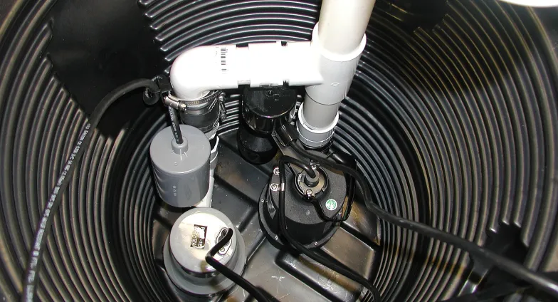 A black sump pump sits in a hole lined with black corrugated plastic. Electrical wires lead to the pump, connected to white PVC pipes rising from the hole.