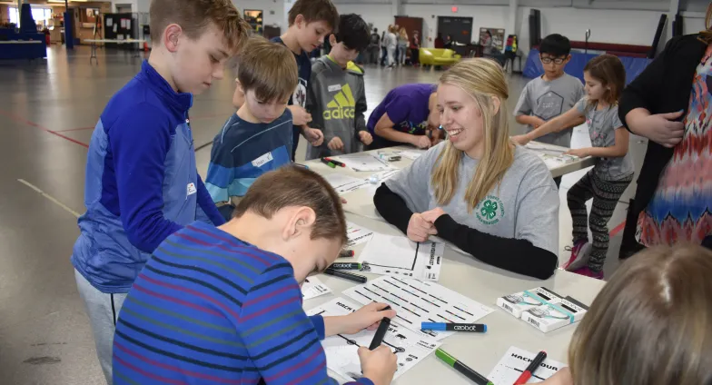 Several kids stand around a curved table, some writing on papers, while a young adult kneels, guiding them.