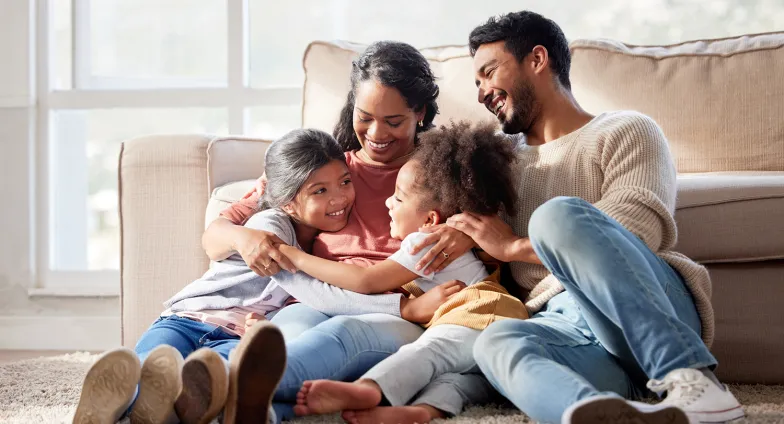 A family of four sits together, hugging, on the floor in front of a beige sofa