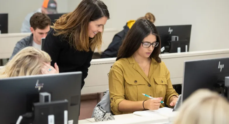 A student sits in front of a computer monitor as a teacher looks over their shoulder.