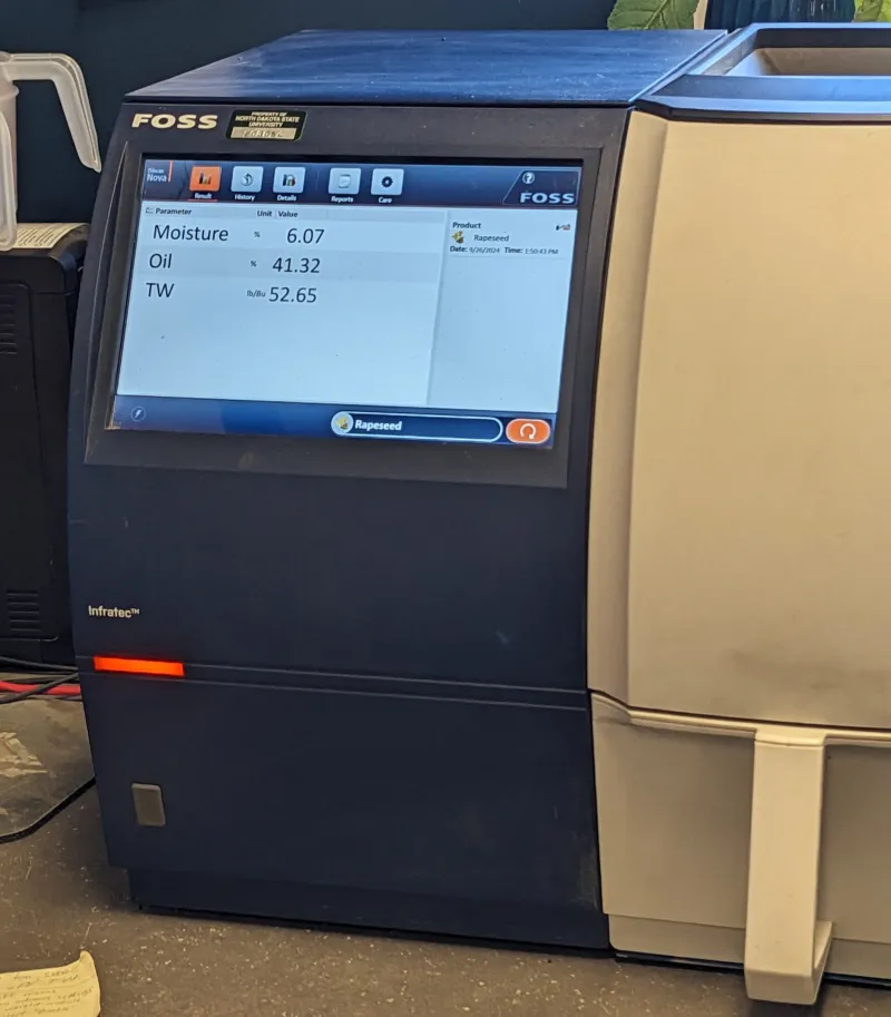 A blue and tan tabletop machine displays moisture, oil, and test weight data on a screen.