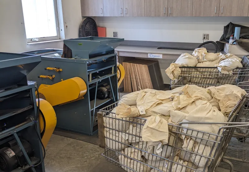 Three wire grocery carts, two of them full of tagged and barcoded cloth bags filled with grain, sit near two small blue and yellow grain cleaners. 