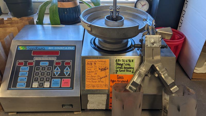 A stainless steel tabletop Seedburo counter has a vibrating metal bowl that feeds kernels across electronics that count each kernel before dumping the seeds into catch basins.  