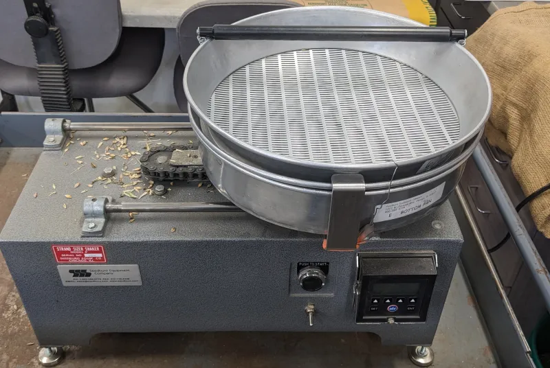 A metal-cased tabletop machine holds a series of slotted screens and a round metal catch basin. A shaker rotates the pans to allow same-sized seed to fall through or remain on top. 