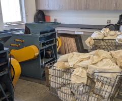 Three wire grocery carts, two of them full of tagged and barcoded cloth bags filled with grain, sit near two small blue and yellow grain cleaners. 