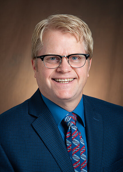 Photo of Dr. Mark Strand with blond hair, wearing glasses, blue suit jacket and tie.