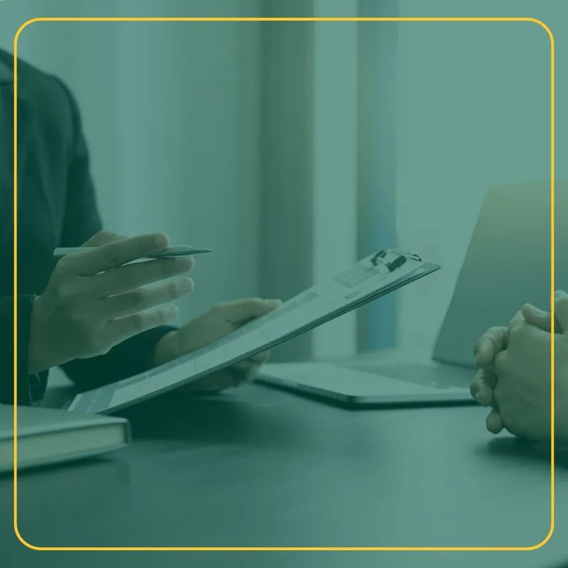Person with a clipboard interviewing someone with hands folded at a desk