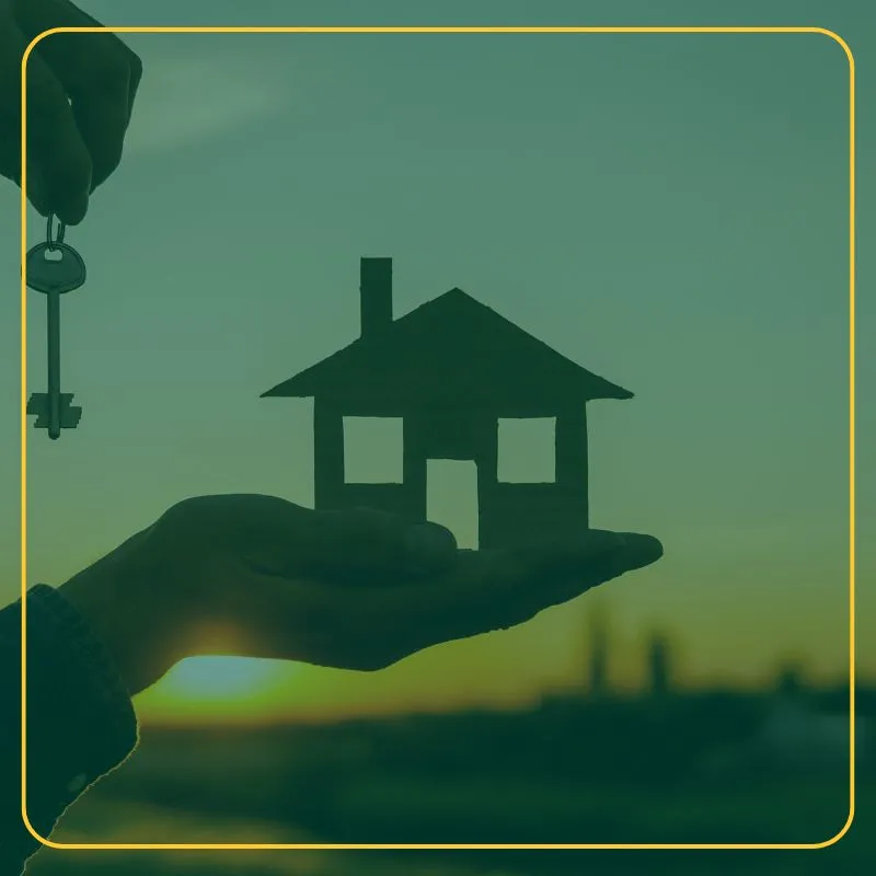 Person holding a model of a house and a key against the backdrop of a sunrise