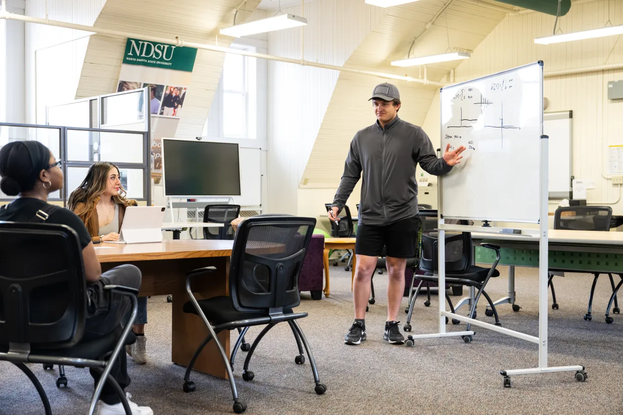 A tutor stands at a whiteboard to demonstrate a mathematics problem.