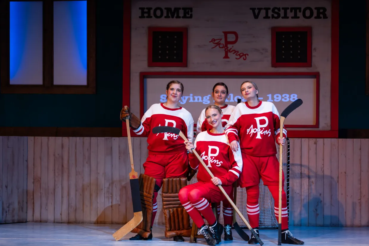 Four female students performing in Theatre NDSU's "Glory." They are wearing red and white hockey uniforms from the 1930s and holding hockey sticks. A scoreboard is behind them.