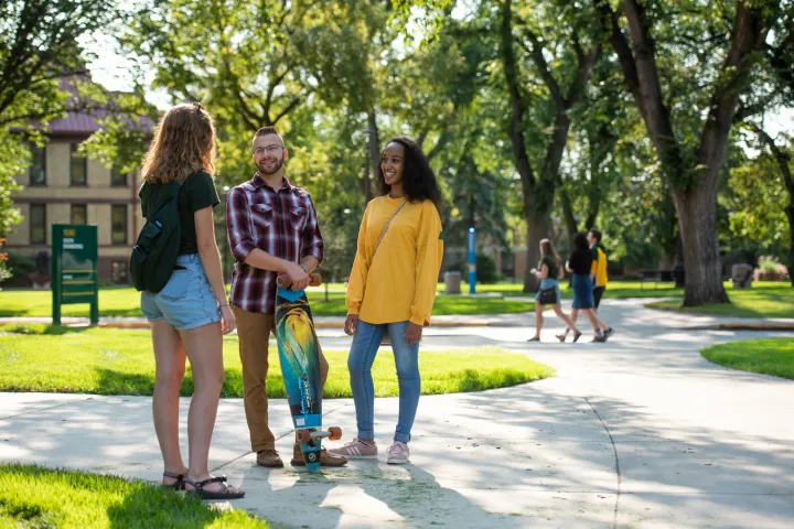 3 NDSU students standing on sidewalk visiting. Background inlcudes green leafy trees, green grass and students walking in the distance.